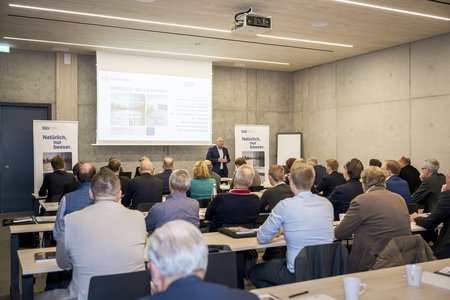 Ein Sprecher hält an einem Podium eine Präsentation vor einem Publikum in einem modernen Konferenzsaal mit Betonwänden.